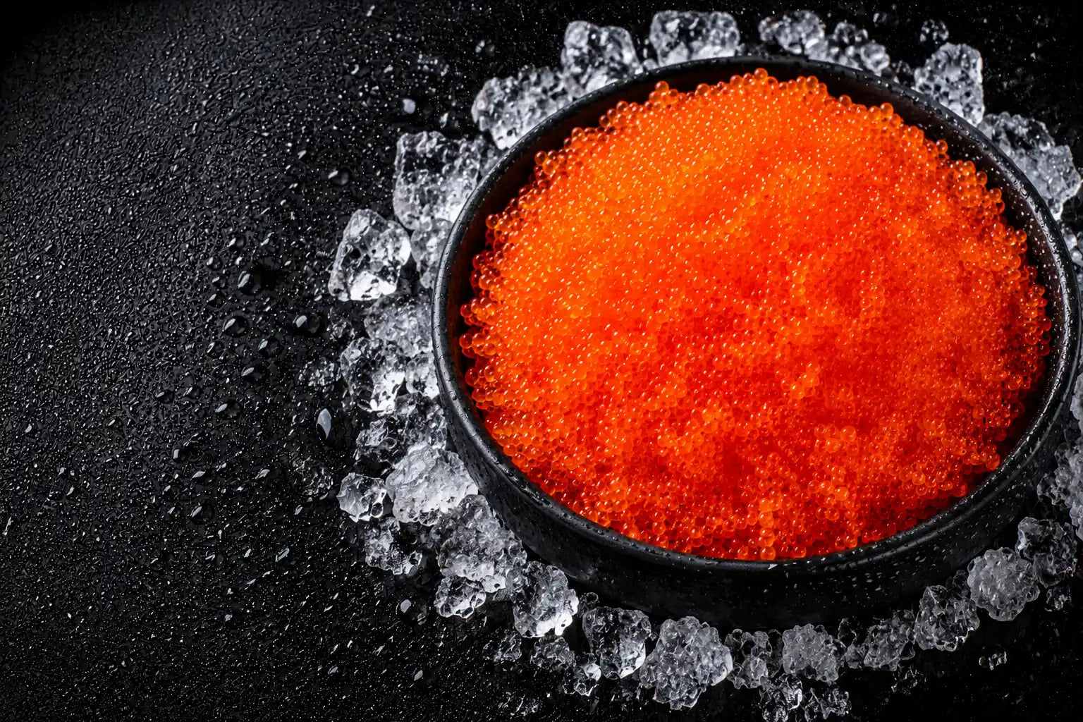 Red caviar in a black bowl surrounded by ice on a dark surface