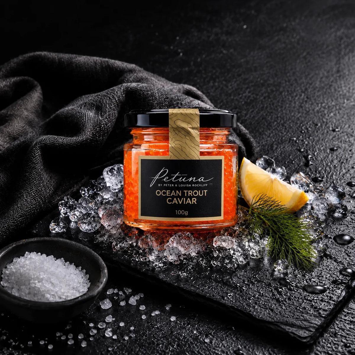 A small glass jar of Petuna Ocean Trout caviar of Tasmania, styled on a dark slate with ice, a lemon wedge, dill, and a small bowl of salt against a dark draped fabric background.