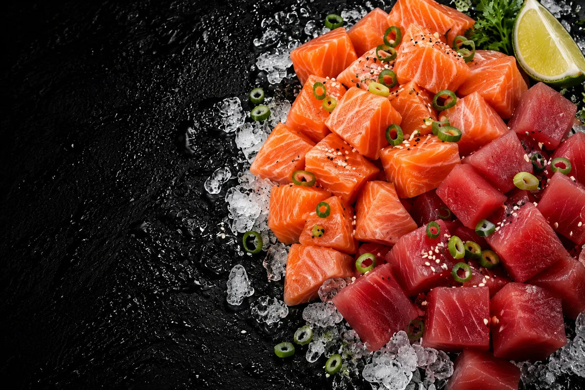 A top-down food photo of raw salmon and tuna sashimi cubes on crushed ice, garnished with sliced green onion and sesame seeds, with a lime wedge in the upper right on a dark textured background.